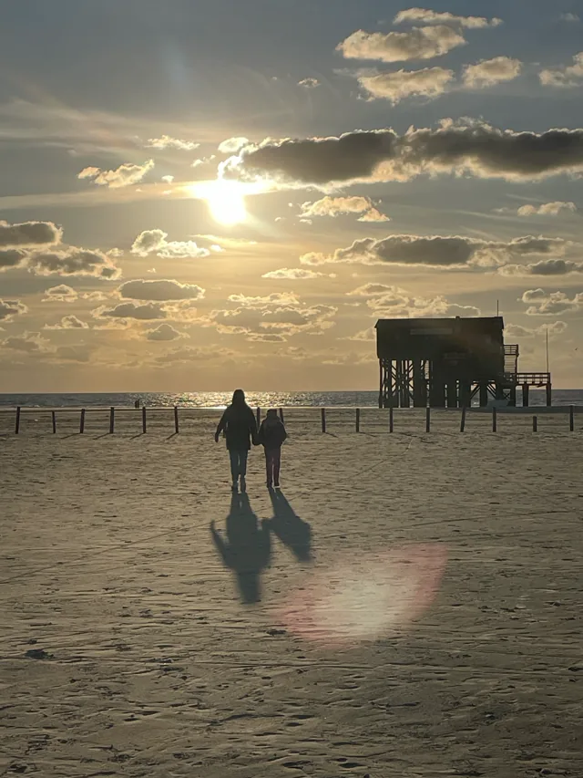 Zwei Menschen gehen am Strand auf das Meer zu, neben einem Pfahlbau im Abendlicht